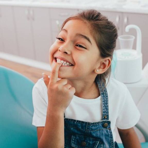 little girl sitting in dentist’s chair and pointing to her teeth 