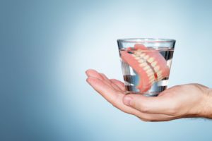 A hand holding up a glass of water with dentures in front of a grayish background