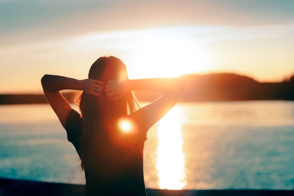 Woman facing sunset at beach with arms behind head